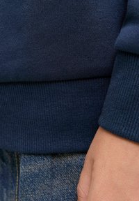 Näheaufnahme einer Hand einer Person, die in der Nähe des Saums eines dunkelblauen Pullovers und einer blauen Jeans ruht.
