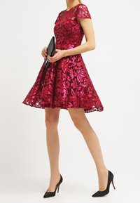 Robe A-line à manches courtes en magenta vif avec superposition en dentelle florale. Associée à une pochette noire et des escarpins pointus noirs.