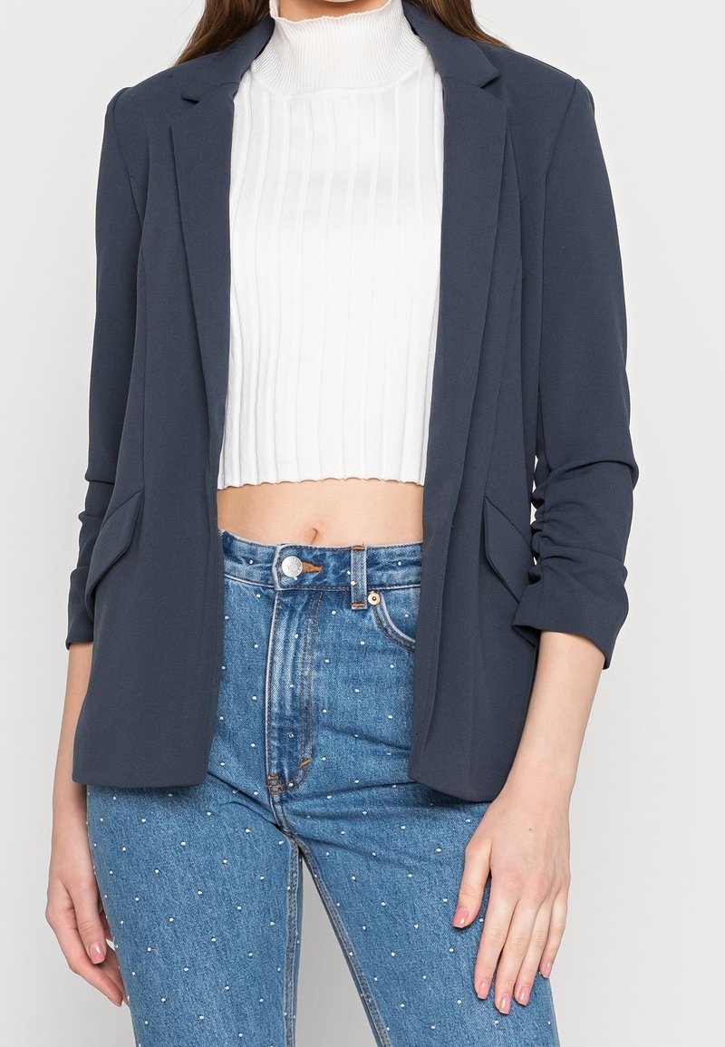 Marineblazer met gestructureerde schouders en open voorkant, gecombineerd met een wit geribbeld crop top, gedragen met een spijkerbroek met hoge taille en versieringen.