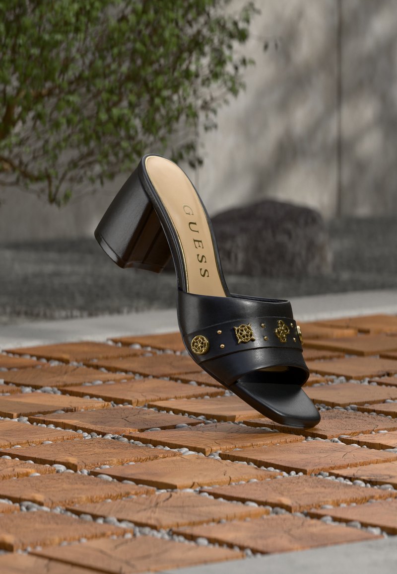 Μαύρο mule με ψηλό τακούνι, διακοσμητικά χρυσά καρφάκια στο λουράκι, τετράγωνη μύτη και λογότυπο "GUESS" στο μπεζ πέλμα, εκτεθειμένο σε ξύλινα πλακάκια έξω.