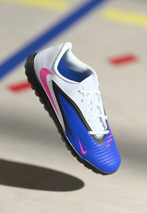 Nogometni čevelj Nike z belim zgornjim delom, modrimi in črnimi poudarki, rožnatim logotipom, teksturiranim dizajnom in gumijastim podplatom. Lahek in zasnovan za gibčnost.