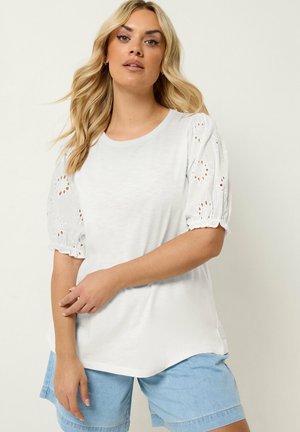 Kobieta z długimi blond włosami, ubrana w biały top z ażurowymi bufkami na rękawach i jasnoniebieskie dżinsowe szorty, stojąca na neutralnym tle.