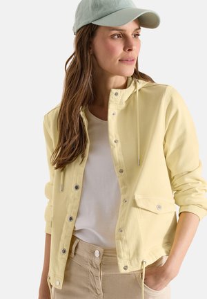 Mujer que lleva una chaqueta amarilla clara abotonada sobre una camisa blanca, pantalones beige y una gorra verde pálido, mirando hacia un lado con una mano en el bolsillo.