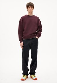 Felpa girocollo bordeaux, vestibilità ampia; jeans in denim neri; scarpe da ginnastica gialle e arancioni con dettagli testurizzati e lacci a contrasto.