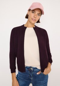 Maglione borgogna con zip, con polsini e colletto a coste, abbinato a una maglietta beige chiaro e jeans in denim. Un berretto in corduroy rosa aggiunge un contrasto di colore.