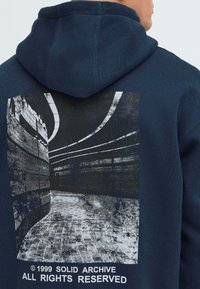 Námornícky modrá mikina s kapucňou, na zadnej strane s grafickým potlačou zakrivenej architektonickej plochy v čierno-bielych farbách a textom nižšie.