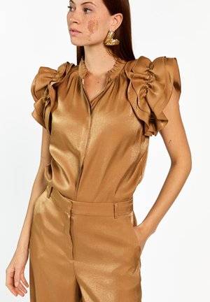 Femme aux longs cheveux bruns et marque de naissance sur le visage, portant un chemisier sans manches à volants en tissu métallique doré et un pantalon taille haute assorti, la main dans la poche.