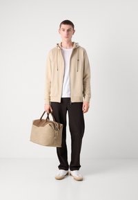Felpa beige con zip sopra una t-shirt bianca, pantaloni neri, sneakers bianche con accenti beige e una borsa a tracolla beige con una strap a contrasto.