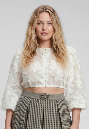 Blonde Frau mit welligem Haar trägt eine weiße, florale, transparente Cropped-Bluse und einen hoch taillierten grauen karierten Rock mit verzierter Gürtelschnalle.