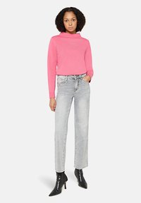 Femme portant un haut à manches longues rose, un jean gris clair et des bottines à talons noires, debout avec une main dans la poche sur fond blanc.