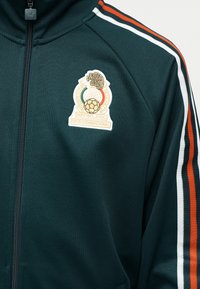 Veste de sport vert foncé avec fermeture éclair intégrale, emblème de l’association de football du Mexique sur la poitrine, et bandes orange, blanche et verte sur la manche.