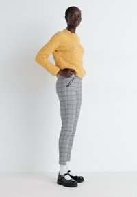 Pull en tricot texturé jaune, pantalon slim en plaid gris avec fermeture éclair sur le côté, chaussettes blanches et chaussures noires à semelle épaisse. Le modèle est présenté de profil.