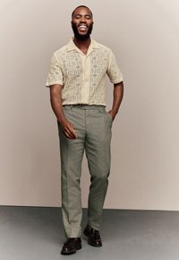 Camicia a maniche corte beige chiaro con texture e dettaglio decorato, abbinata a pantaloni grigio chiaro e scarpe scure, su un pavimento grigio.