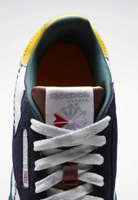 Reebok joggesko med en hvit mesh-overdel, marineblå semsket skinn-elementer, grønne og gule detaljer, og hvite snørebånd. Har en logomerket etiket på tungen.
