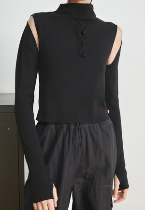 Haut noir à manches longues avec épaules dénudées, associé à un pantalon noir ample, porté par une personne debout à l'intérieur.