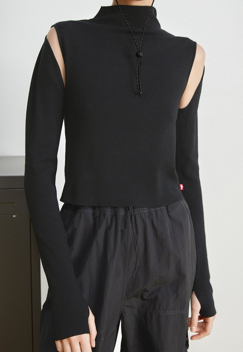 Haut noir à manches longues avec épaules dénudées, associé à un pantalon noir ample, porté par une personne debout à l'intérieur.
