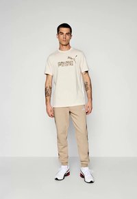 T-shirt de algodão bege com um logo camuflado "PUMA", emparelhada com calças de treino bege e sapatilhas brancas com detalhes em preto e vermelho.