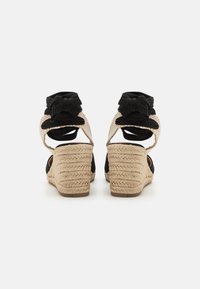 Sorte kile-sandaler i svart med en flettet jute-helspreng, åpen bak og stoffblomsterdetaljer ved ankelen. Teksturerte overflater og justerbare stropper.