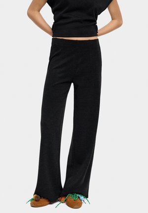 Kobieta ubrana w czarne, szerokie spodnie i czarny top bez rękawów oraz brązowe buty z kolorowymi sznurowadłami i pomponami.