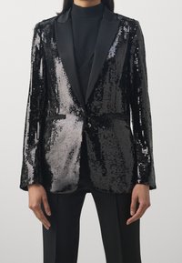 Blazer negro con lentejuelas y solapas de satén; corte ajustado con bolsillos delanteros y una superficie texturizada y brillante que refleja la luz.