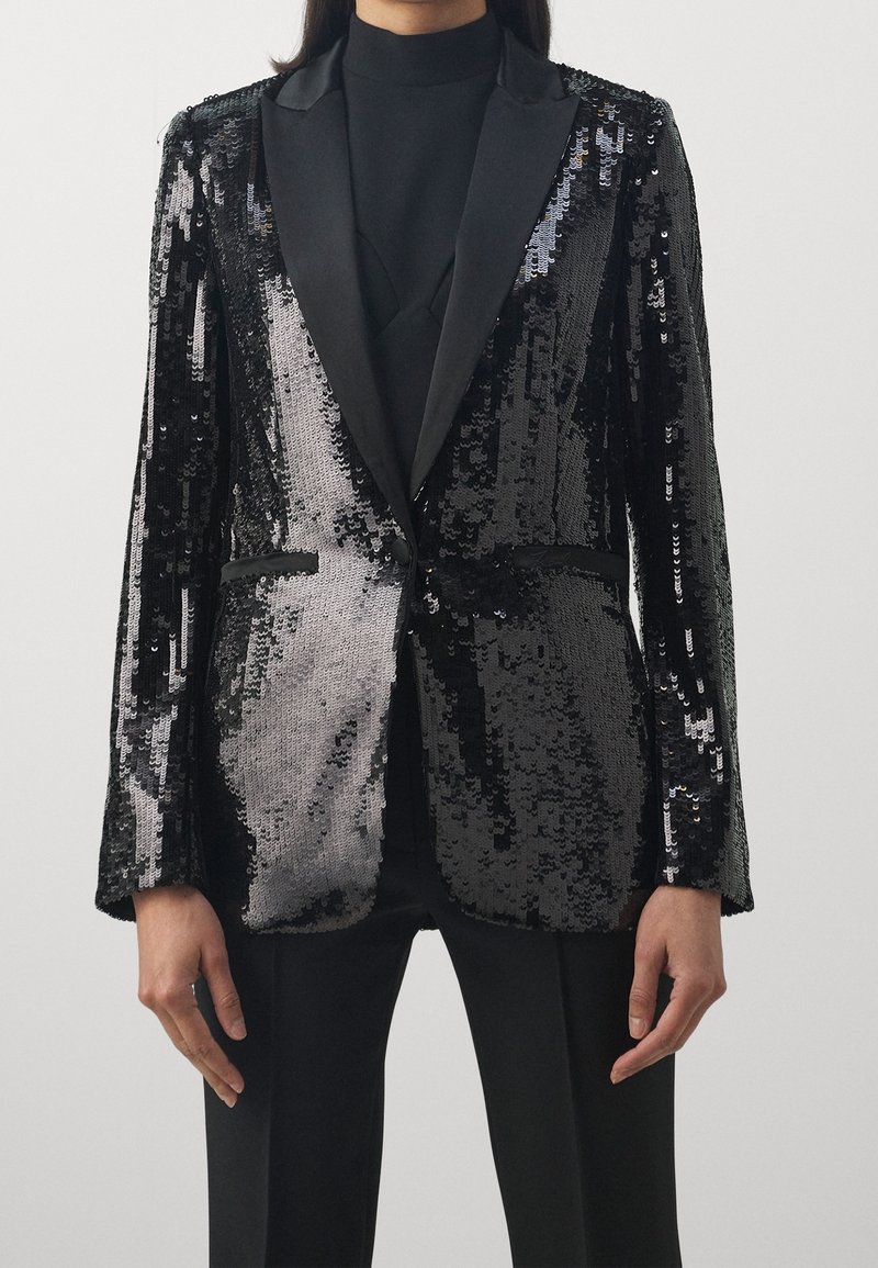 Blazer negro con lentejuelas y solapas de satén; corte ajustado con bolsillos delanteros y una superficie texturizada y brillante que refleja la luz.