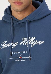 Hanorac albastru din bumbac, cu text brodat alb "Tommy Hilfiger", șnururi cu vârfuri argintii și un mic detaliu cu logo.