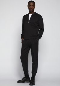 Chaqueta bomber negra con cremallera combinada con pantalones tapered a juego. Tejido suave con puños acanalados y una camiseta blanca de cuello redondo debajo.