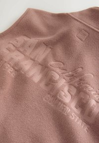 Fleece-Weste in sanftem Rosa mit geprägtem Text "SAN FRANCISCO". Verfügt über einen runden Kragen und eine subtile Textur. Sauberes, minimalistisches Design mit Logodetail.