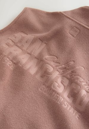 Fleece vest in soft pink with embossed "SAN FRANCISCO" text. Features a rounded collar and subtle texture. Clean, minimalist design with logo detail.