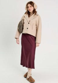 Veste en mélange de laine beige à double boutonnage, jupe midi satin bordeaux, mules en fourrure marron et sac à main en imprimé léopard. Des cache-oreilles complètent le look.