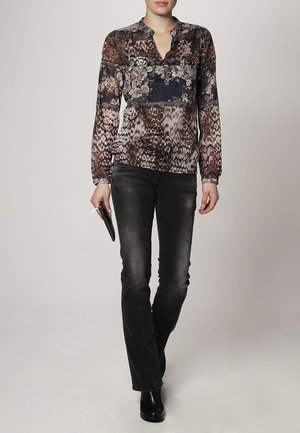 Kvinde iført en mønstret langærmet bluse, sorte vasket jeans, sorte støvler, holder en lille sort clutch.