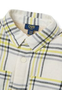 Camisa de manga larga con cuello en color crema con un patrón de cuadros en gris, amarillo y negro; cuenta con dos bolsillos frontales y cierres de botón blancos.