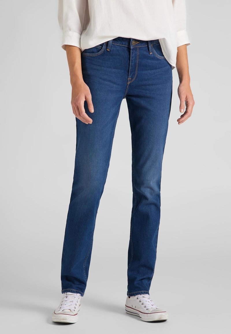 Personne portant un jean bleu slim, une chemise blanche à manches longues et des baskets blanches debout sur un fond gris.