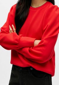 Roter Pullover mit langen Ärmeln, gerafften Bündchen und Rundhalsausschnitt. Kombiniert mit schwarzen, hochtaillierten Jeans. Glatte Stofftextur.