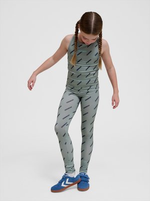 Young girl with braided hair wearing gray sportswear printed with "HUMMEL" and blue sneakers, standing and looking down.