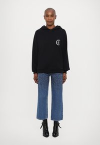 Felpa nera con cappuccio e logo frontale, abbinata a jeans blu a gamba larga con motivo testurizzato e stivali neri alla caviglia.