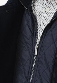 Podszewka pikowanej granatowej kurtki z srebnym zamkiem błyskawicznym i czarną warstwą zewnętrzną, zestawiona z białą koszulą w granatową kratę.