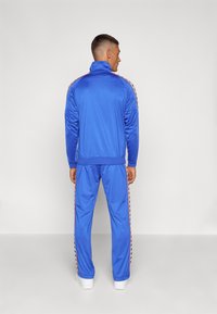 Hemelsblauw trainingspak met een hoge kraag, lange mouwen en zijstrepen met een logo. Gemaakt van een gladde, lichte stof.