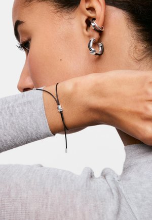 Primer plano de la oreja de una persona con dos pendientes de aro plateados y negros y una muñeca con una pulsera negra y fina con un pequeño colgante plateado.
