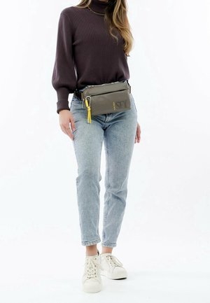 Borsa a cintura in tessuto grigio con superficie strutturata, dotata di un pon-pon giallo e chiusura con zip. Indossata sopra jeans azzurri chiari e sneakers bianche.