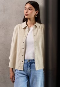 Lichtbeige corduroy knoopshirt met een kraag, gedragen over een wit T-shirt en gecombineerd met lichtblauwe jeans.