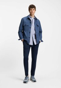 Mężczyzna ubrany w niebieską zapinaną na guziki kurtkę na jasnoniebieską koszulę, ciemnogranatowe spodnie i jasnoniebieskie trampki, stojący z rękami w kieszeniach.