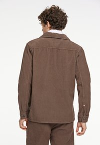 Chaqueta de pana marrón con cuello de solapa, mangas largas y puños abotonados; presenta una textura de cordones verticales y un corte relajado.