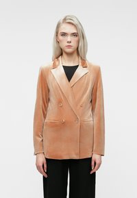 Femme blonde portant un blazer double boutonnage en velours pêche sur un haut noir et un pantalon noir, se tenant devant un fond blanc uni.