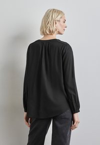 Blusa nera a maniche lunghe con collo rotondo, caratterizzata da dettagli a pieghe sulle spalle, tessuto morbido e orlo curvo. Abbinata a pantaloni grigi.