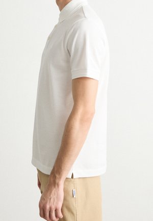 Mann trägt ein weißes kurzärmeliges Polohemd und beige Hose, von der Seite gezeigt mit locker herabhängendem Arm.