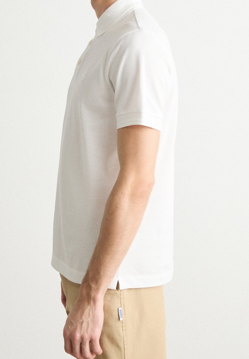 Homme portant un polo blanc à manches courtes et un pantalon beige, montré de profil avec le bras détendu vers le bas.