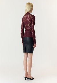 Femme aux longs cheveux blonds porte un haut en dentelle bordeaux à manches longues, une jupe en cuir noire fendue à l'arrière, et des mules noires à talons bas, debout de dos.
