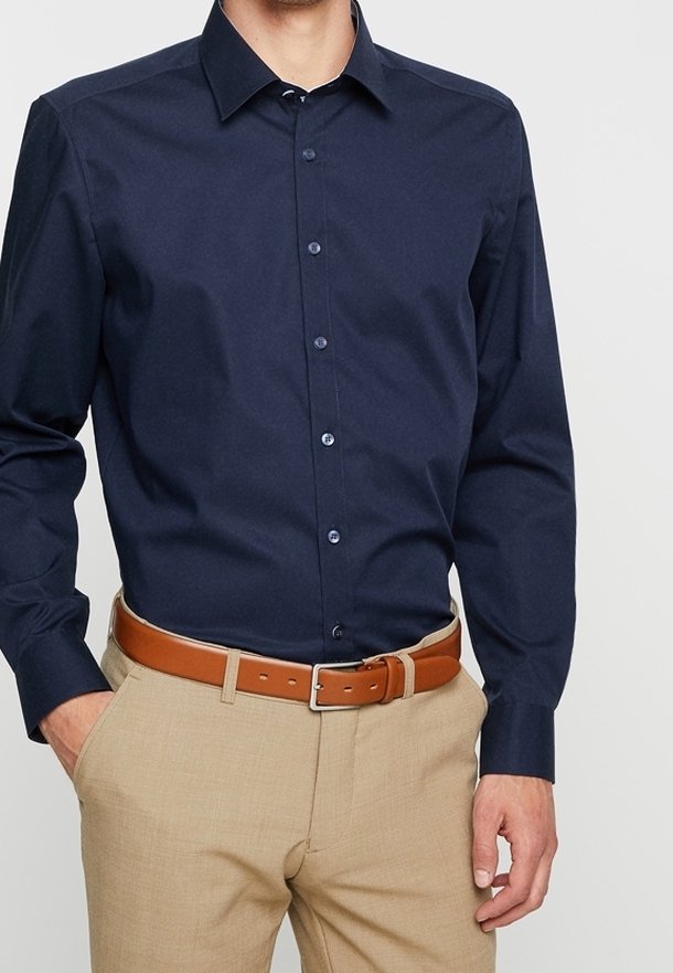 Homme portant une chemise bleu foncé à manches longues boutonnée rentrée dans un pantalon beige avec une ceinture marron, mains partiellement dans les poches.