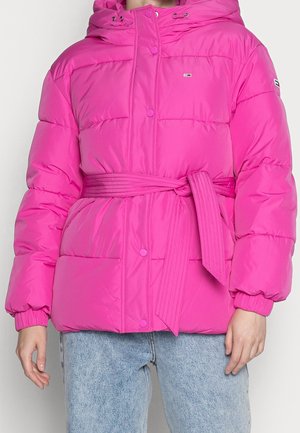 Lyserød dunjakke med hætte, knaplukning foran og matchende bredt bælte bundet i taljen, båret med lyseblå jeans.